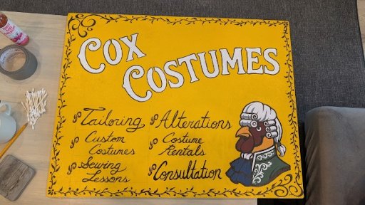 The hand-painted Cox Costumes shop sign: white lettering on mustard yellow, a vine border, and a rooster in a powdered wig and blue coat.